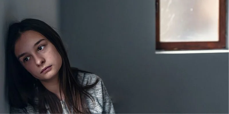 A sad teenage girl with dark hair leans against a wall