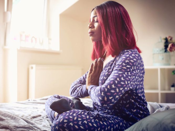 An African American woman sits on her bed and meditates to manage anxiety