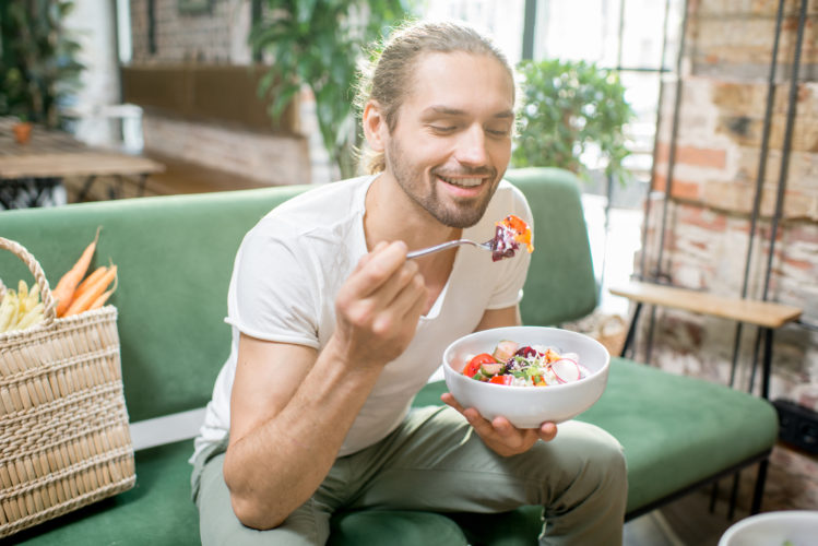 Handsome man eating healthy salad sitting indoors on the green sofa with bag full of vegetables on the background. Healthy eating concept