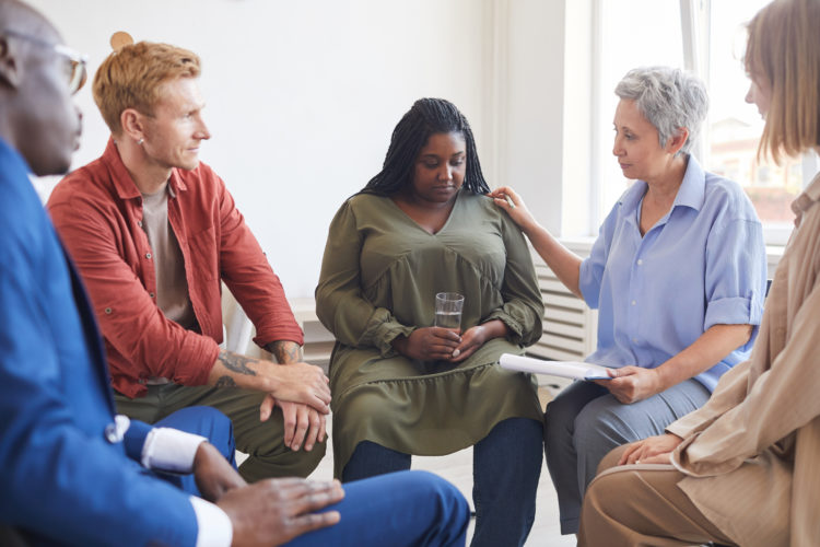 Portrait of young African-American woman sharing struggles during support group meeting with people siting in circle and comforting her