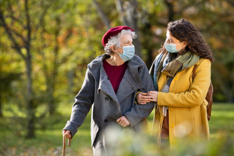 Lovely granddaughter walking with senior woman holding stick in park and wearing mask for safety against covid-19. Happy old grandmother enjoying walking in park with girl. Smiling elderly woman with happy caregiver in park relaxing after quarantine due to coronavirus outbreak and lockdown.