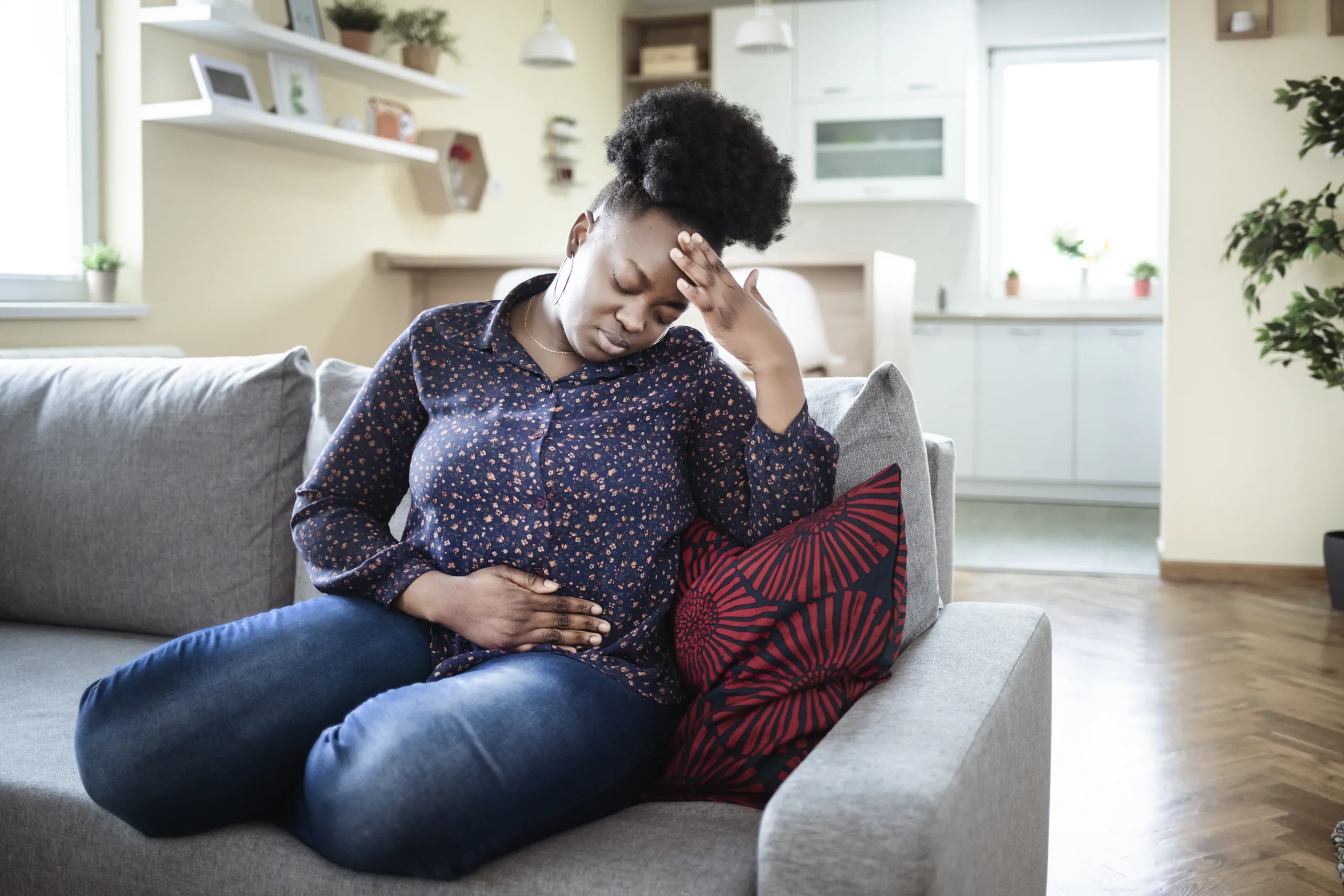 Young sick african american plus size woman with hands holding pressing her crotch lower abdomen. Medical or gynecological problems, healthcare concept. Young woman suffering from abdominal pain while sitting on sofa at home