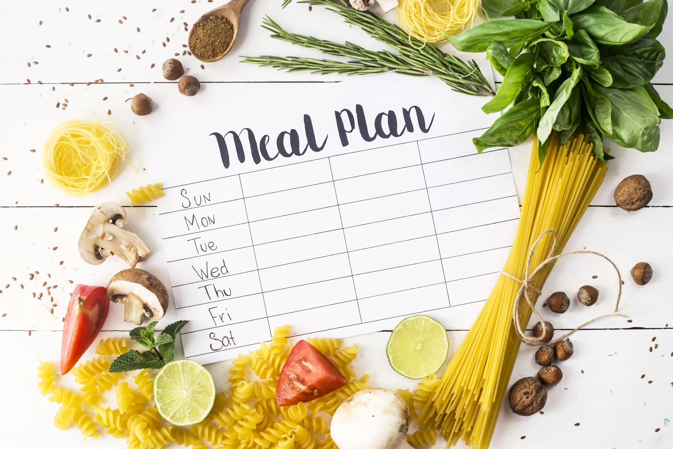 A meal plan for a week on a white table among products for cooking - pastas, basil, vegetables, lime, seeds, nuts and spices. Top view, flat lay, copyspace