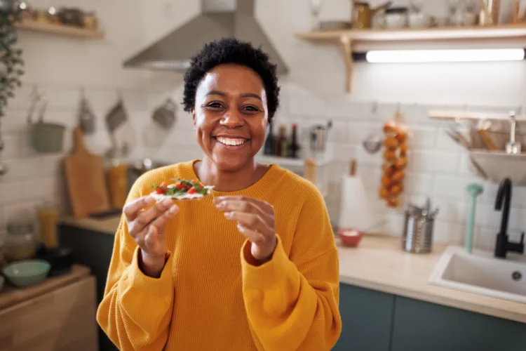 Portrait of young smiling beautiful woman in a domestic kitchen, holding cracker with vegetables in her hand