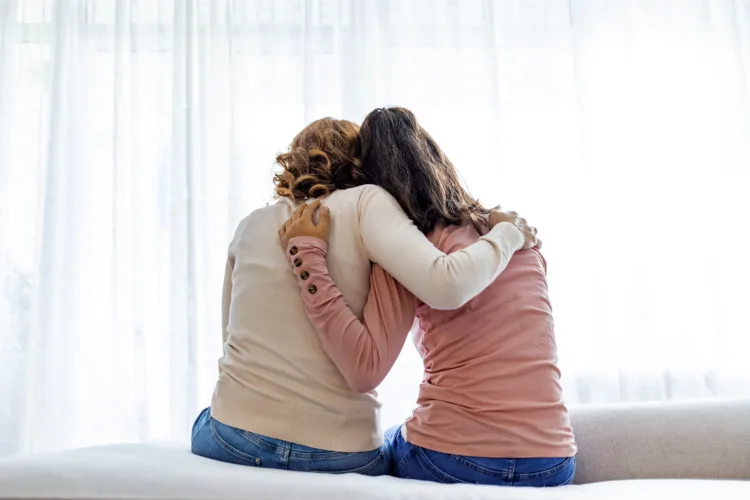 Rear back view of a mother and daughter embrace sitting on bed at home, older sister consoling younger teen, girl suffers from unrequited love share secrets trustworthy person relative people concept