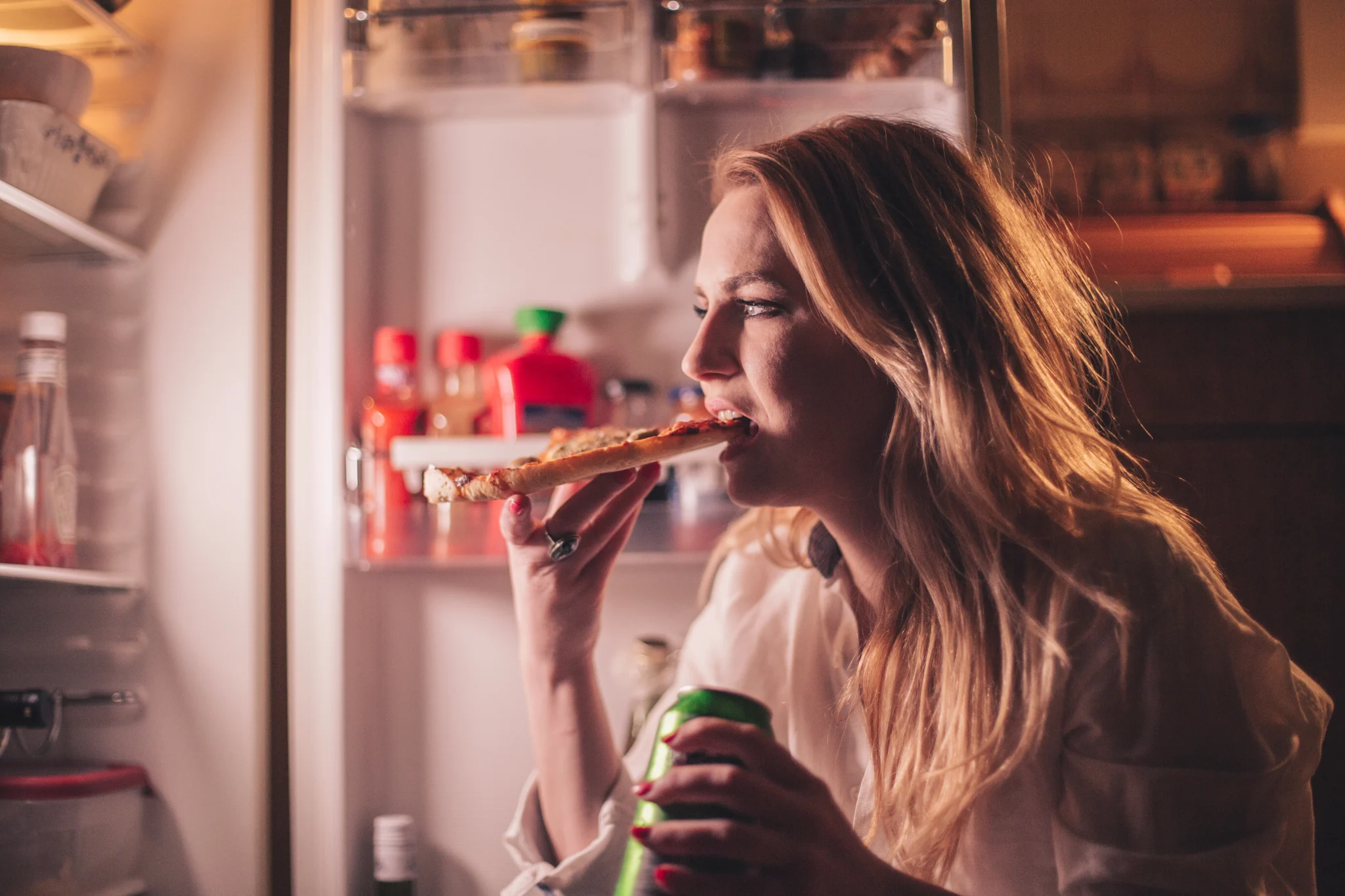 Young woman binge eating pizza slice and drinking beer during the night