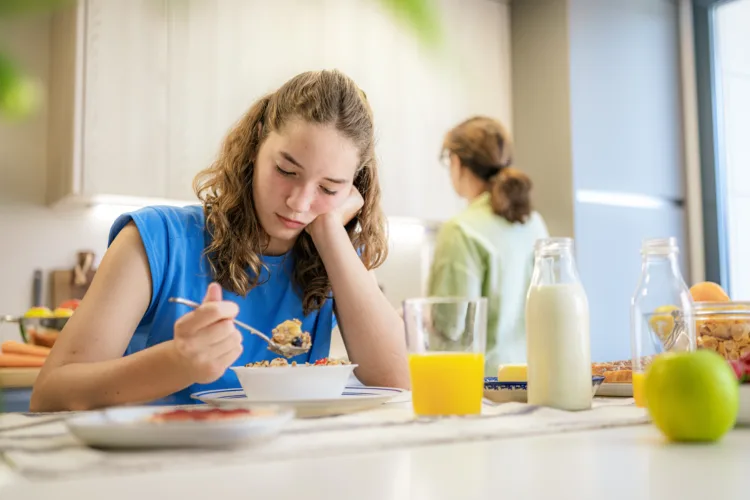 Upset and distraught teen girl eating breakfast in the kitchen while her mother is doing chores